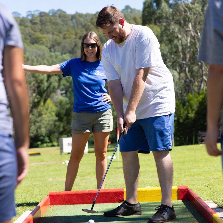 Participant playing mini golf outdoors with a support worker nearby